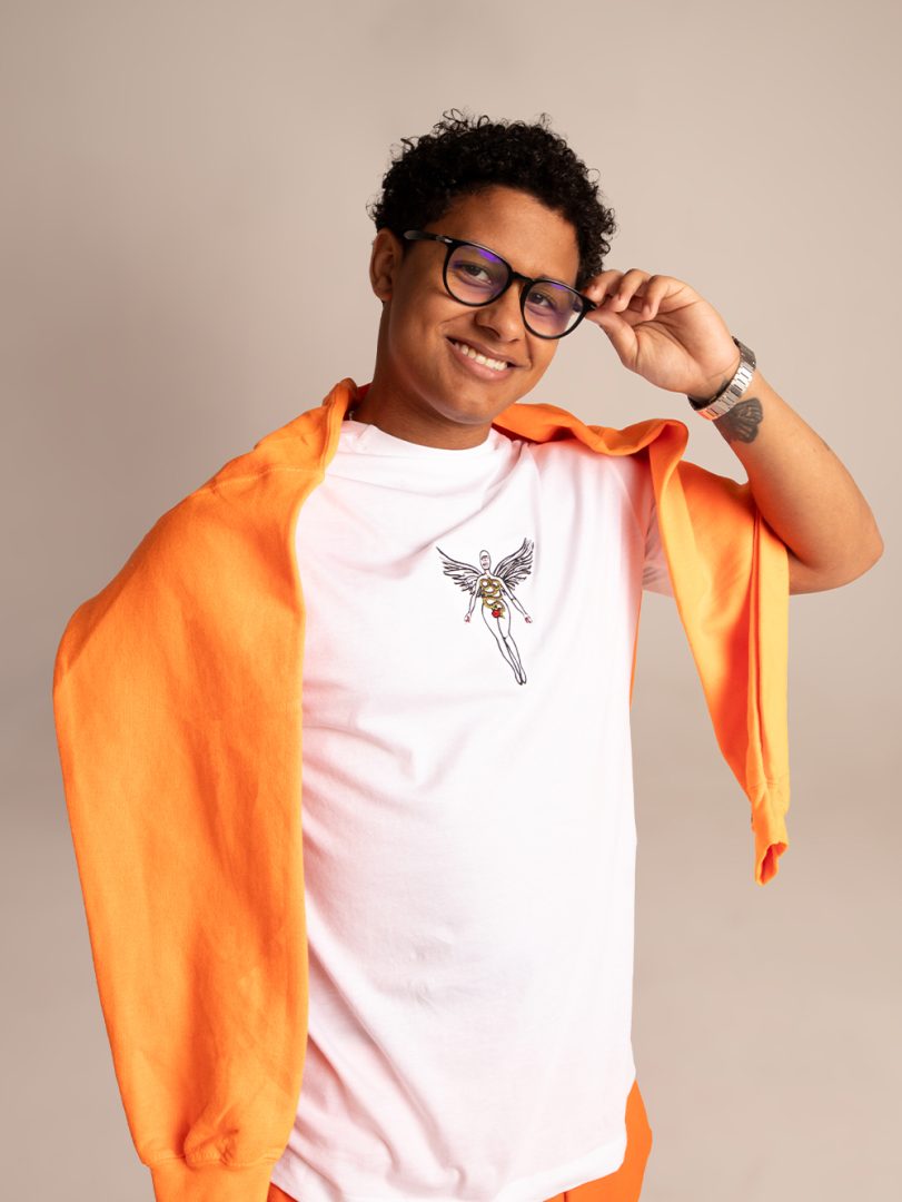 A smiling model poses against a beige background wearing a white Checkmate graphic tee with an orange sweatshirt draped around the neck.