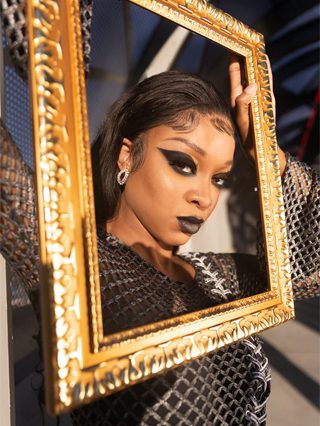 An editorial-style photo of a model wearing the Checkmate Anywear Nia Long Metallic Dress with a gold-detailed face frame, set against a softly blurred landscape background.