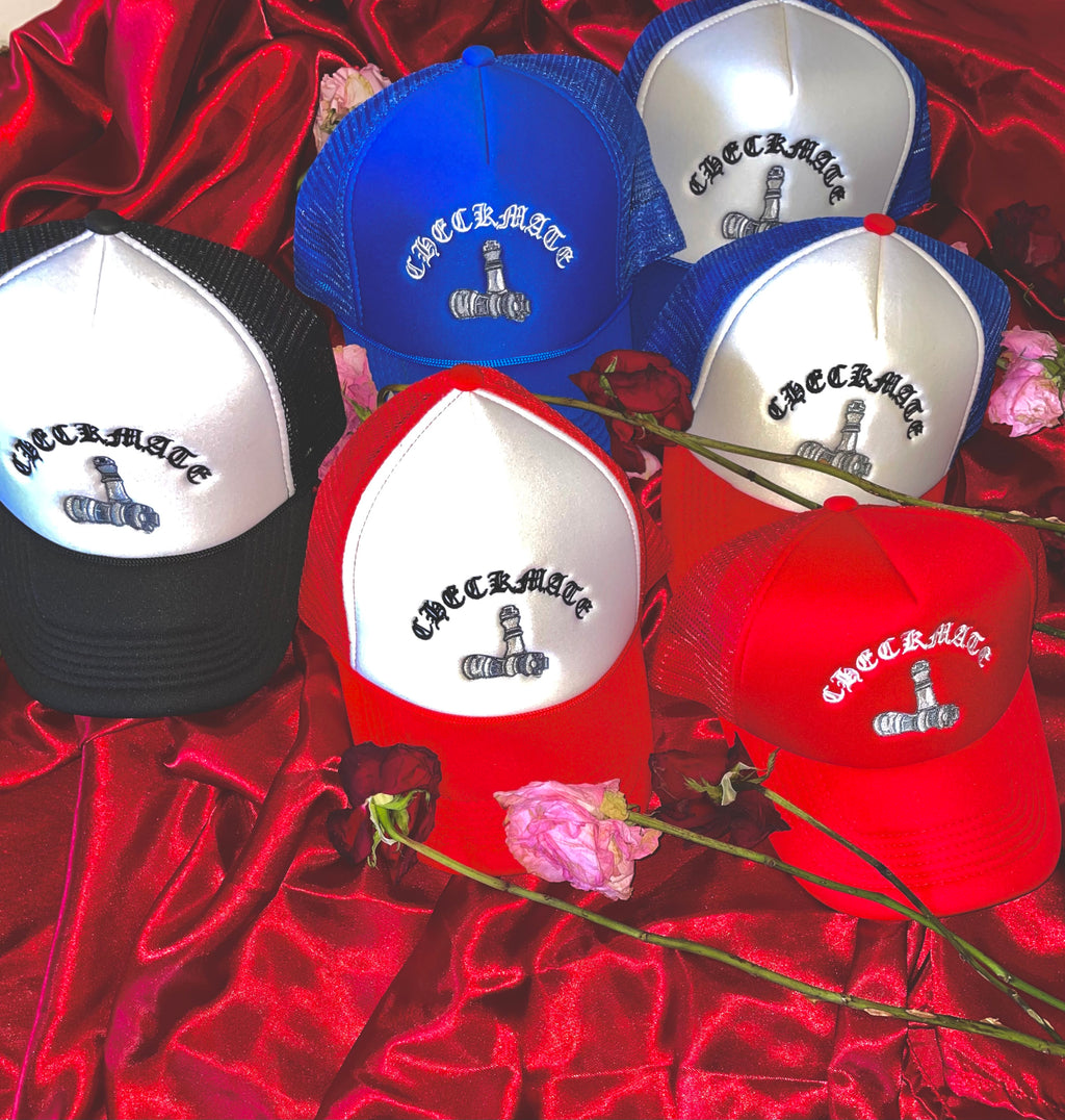 A group of Checkmate Anywear trucker hats in red, blue, black, and white are displayed on a red satin background. The hats are arranged to highlight their bold colors and mesh-back design.