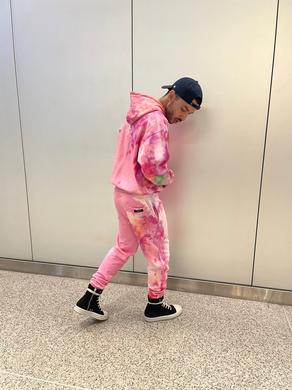 A model is shown from the back wearing a pink tie-dye sweatsuit by Checkmate Anywear, paired with a hat and sneakers. The outfit has a relaxed, streetwear-inspired fit.