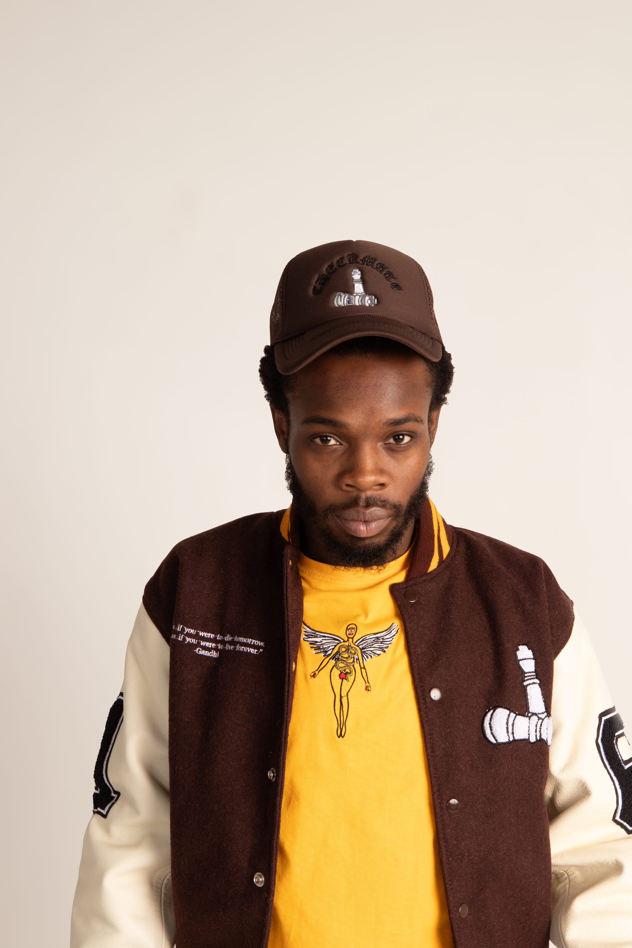 Man wearing a brown jacket, yellow shirt with graphic design, and brown cap on a plain background
