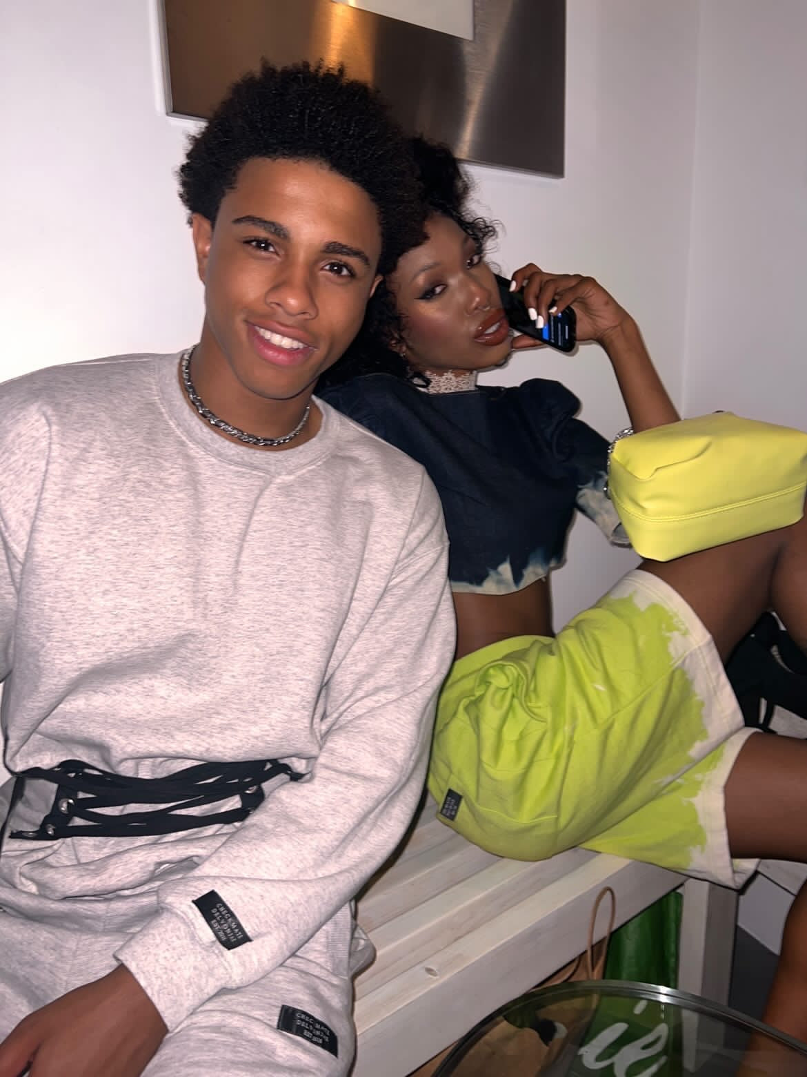 Two people sitting together, one using a phone, in a casual indoor setting.