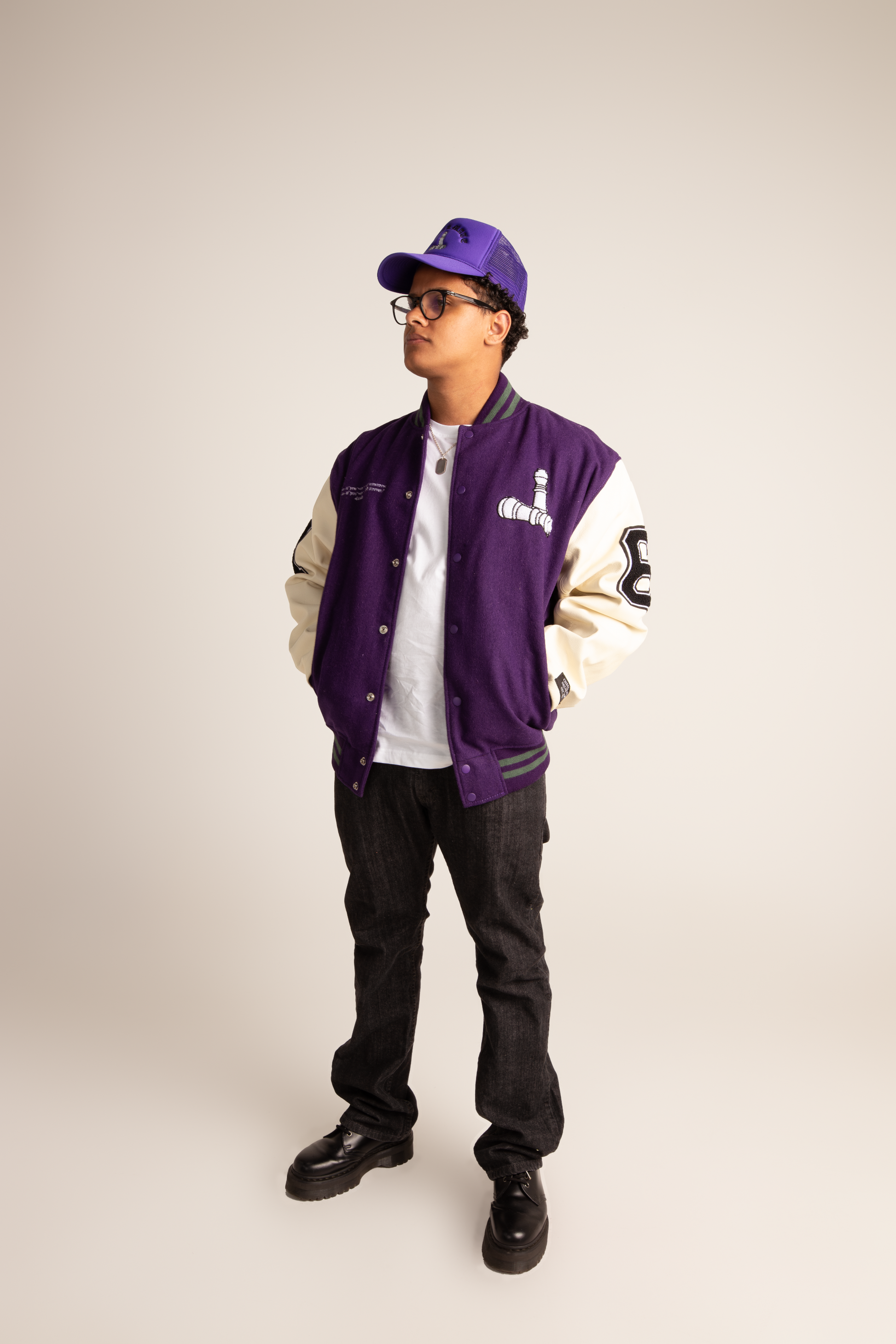 Man wearing a checkmate anywear varsity jacket with white sleeves, white shirt, black pants, and a purple  checkmate anywear cap on a netural background.
