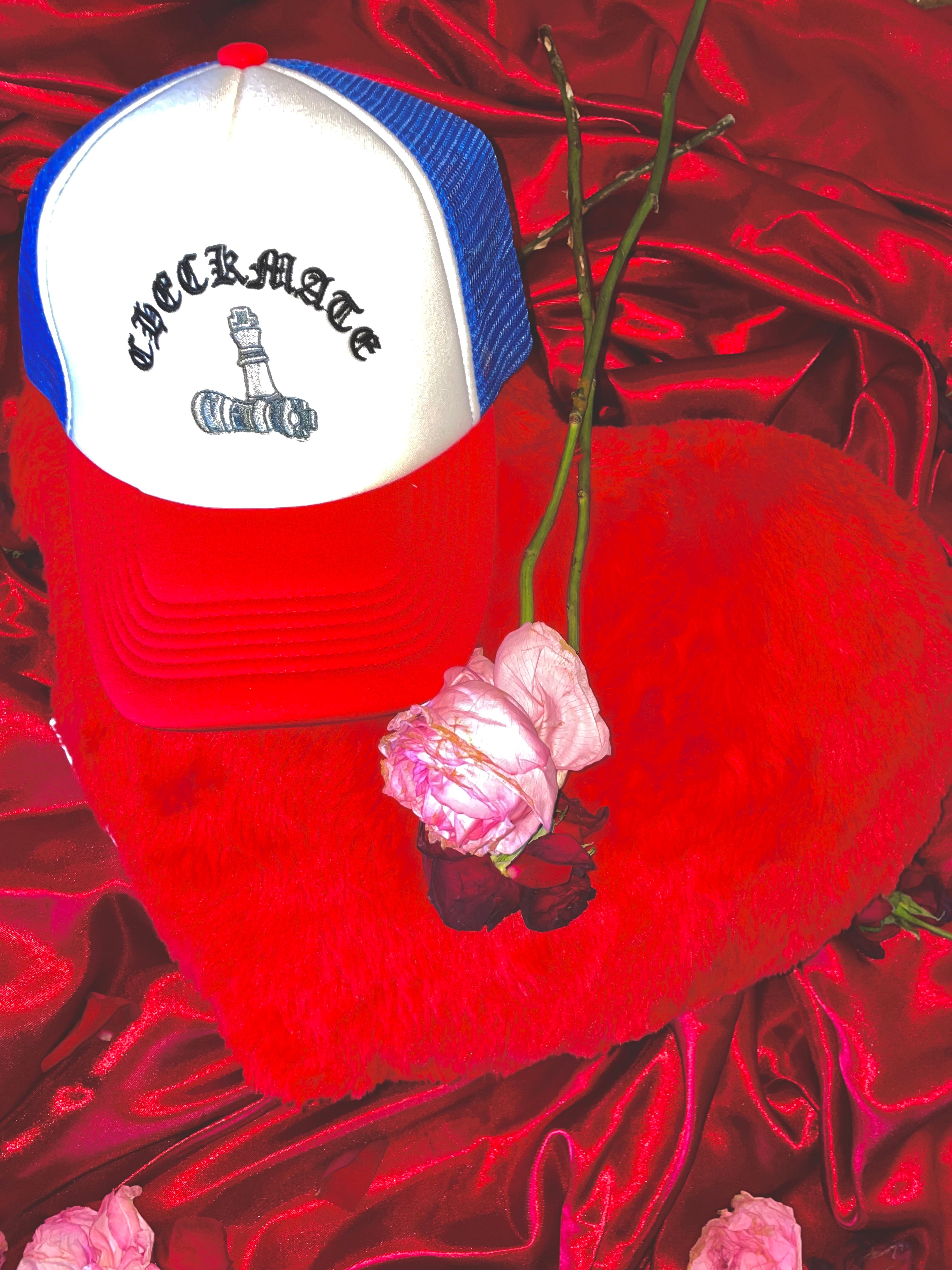 Red. Blue, and white checkmate anywear trucker hat on heart-shaped pillow with a pink flower on a red satin background